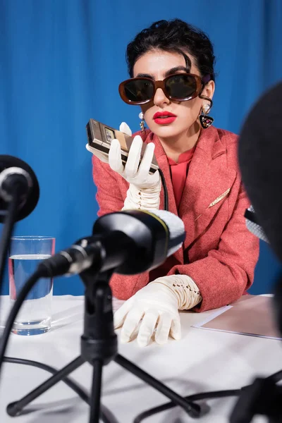 Blurred microphones near retro style woman holding voice recorder during interview on blue background — Stock Photo