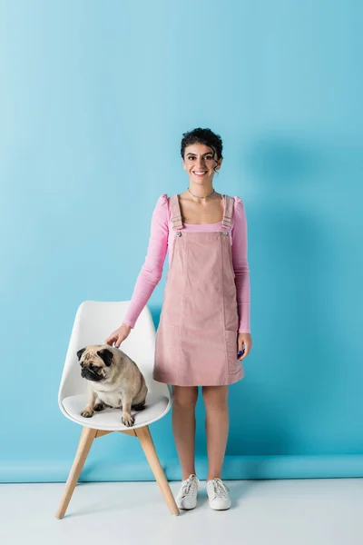 Full length view of smiling woman in pastel clothes near pug on chair on blue background — Stock Photo