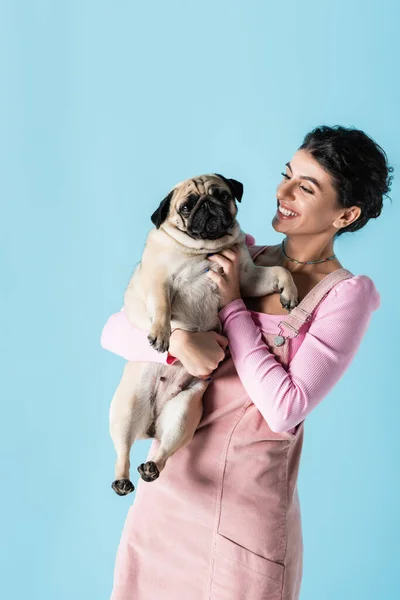 Junge und fröhliche Frau kuschelt Mops Hund, während sie isoliert auf blau steht — Stockfoto