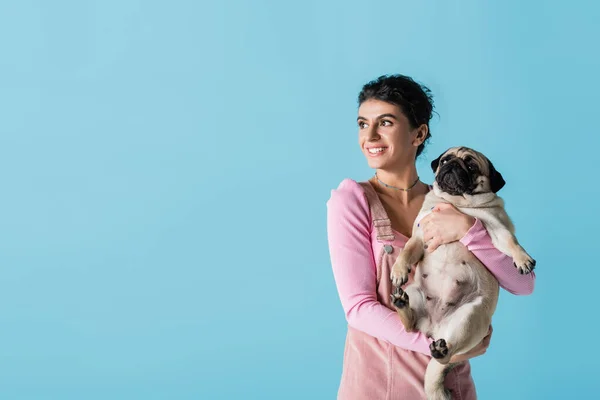 Mujer complacida mirando hacia otro lado mientras sostiene perro pug aislado en azul - foto de stock