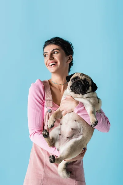 Donna sorridente distogliendo lo sguardo e tenendo cane carlino isolato su blu — Foto stock