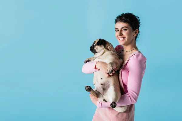 Young and trendy woman holding pug dog while smiling at camera isolated on blue — Stock Photo