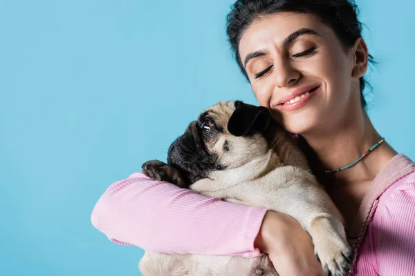 Glückliche brünette Frau mit geschlossenen Augen umarmt Mops Hund isoliert auf blau — Stockfoto