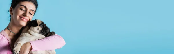Felice donna bruna con gli occhi chiusi abbracciando cane carlino isolato su blu, banner — Foto stock