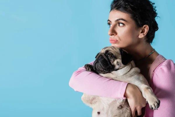 Pensativo morena mujer abrazando pug perro mientras mira lejos aislado en azul - foto de stock