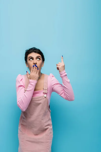 Mujer conmocionada mirando hacia arriba, señalando con el dedo y cubriendo la boca con la mano aislada en azul — Stock Photo