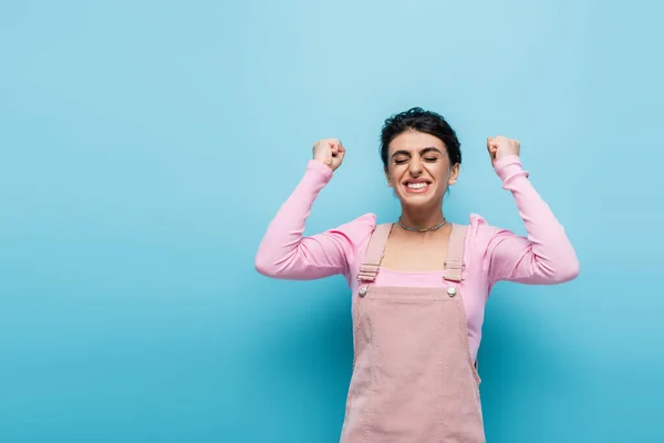 Mujer llena de alegría mostrando sí gesto mientras está de pie con los ojos cerrados aislados en azul - foto de stock