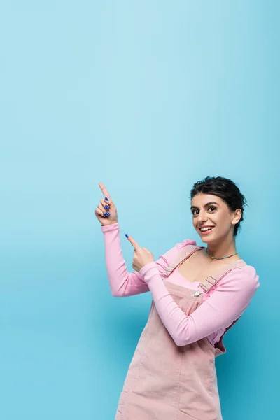 Young brunette woman in pastel clothing pointing with fingers and looking at camera isolated on blue — Stock Photo