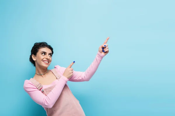 Jovem alegre em roupas pastel na moda olhando para longe e apontando com os dedos isolados no azul — Fotografia de Stock