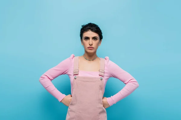 Serious young woman in pastel clothes standing akimbo on blue background — Stock Photo