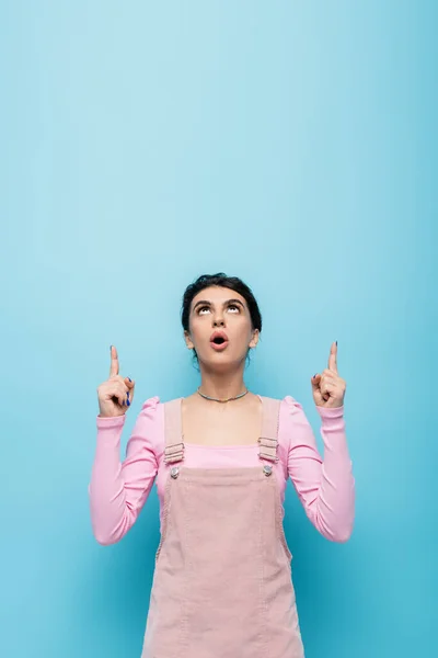 Astonished woman in pastel clothing looking up and pointing with fingers isolated on blue — Stock Photo