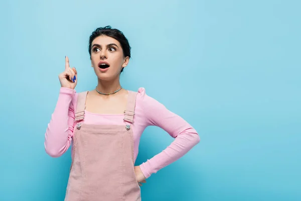 Mujer emocionada en ropa pastel de moda de pie con la mano en la cadera y mostrando gesto idea aislado en azul - foto de stock