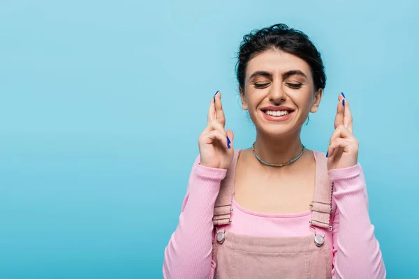 Woman with closed eyes and crossed fingers smiling isolated on blue — Stock Photo