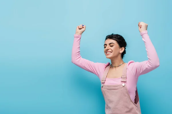 Mujer alegre y elegante con los ojos cerrados mostrando gesto de triunfo sobre fondo azul - foto de stock