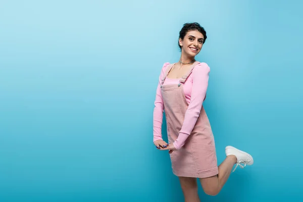 Happy and coquettish woman looking at camera while standing on one leg on blue background — Stock Photo