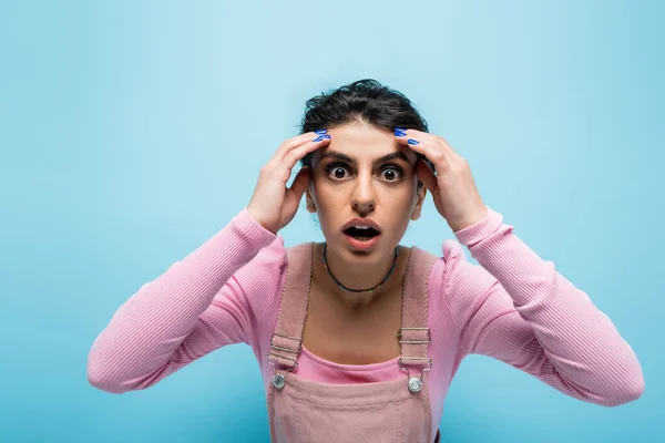 Shocked brunette woman touching head and looking at camera isolated on blue — Stock Photo