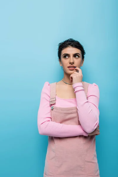 Mujer de ensueño en ropa pastel tocando el labio mientras mira hacia otro lado aislado en azul - foto de stock