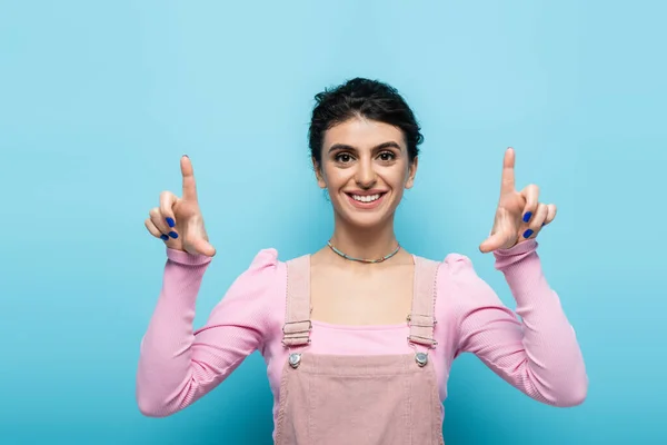 Vista frontal de la mujer alegre y elegante apuntando hacia arriba con los dedos aislados en azul - foto de stock