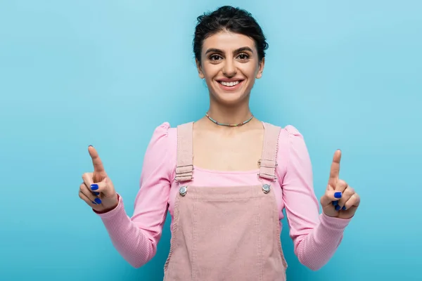 Vista frontal de la mujer alegre apuntando hacia arriba con los dedos aislados en azul - foto de stock