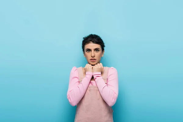 Worried woman holding fists near chin while looking at camera isolated on blue — Stock Photo