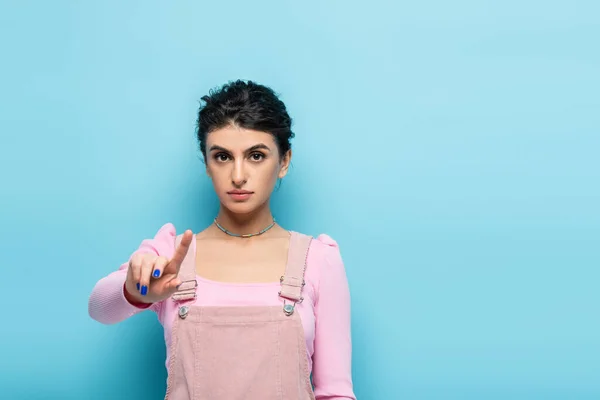 Junge brünette Frau zeigt mit dem Finger und schaut in die Kamera auf blauem Hintergrund — Stockfoto