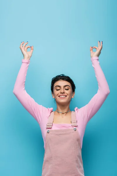 Усміхнена жінка з закритими очима, роздумуючи з піднятими руками і жестом Джана Мудра ізольовані на синьому — стокове фото