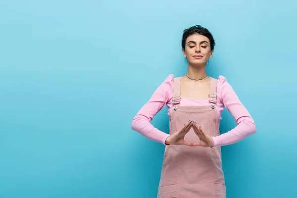 Mujer positiva en ropa pastel relajante en la meditación pose aislada en azul - foto de stock