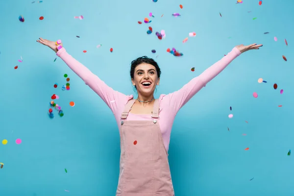 Amazed and happy woman with raised hands under falling confetti on blue background — Stock Photo