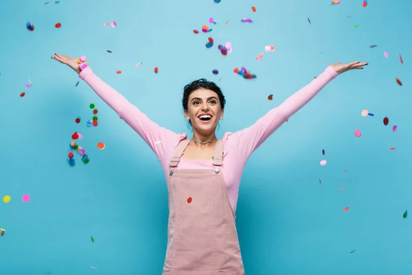 Mulher espantada e feliz com as mãos levantadas sob queda confetti no fundo azul — Fotografia de Stock