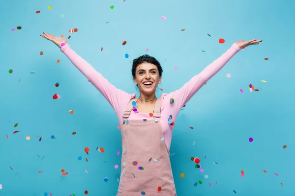 Excited woman in pastel clothes smiling under falling confetti on blue background — Stock Photo
