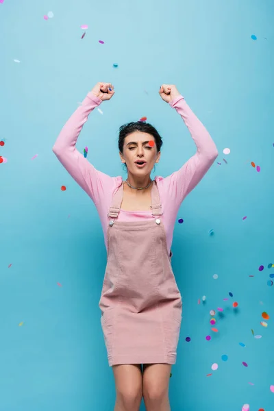 Excited woman with closed eyes showing triumph gesture under falling confetti on blue background — Stock Photo