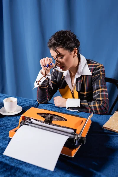 Periodista cansado sentado con los ojos cerrados cerca de taza de café y máquina de escribir vintage sobre fondo azul - foto de stock