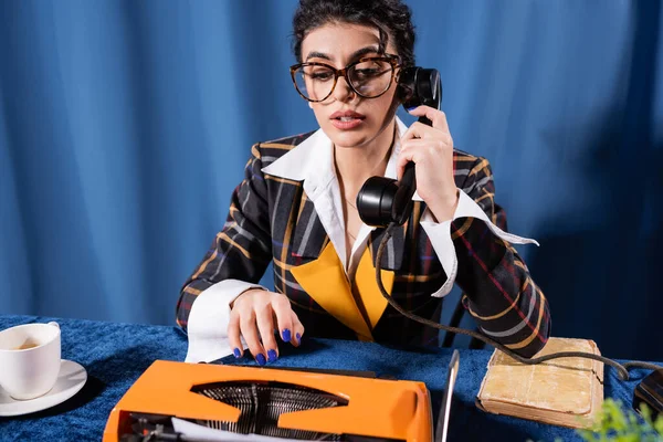 Periodista morena en chaqueta de estilo retro y anteojos hablando por teléfono cerca de la máquina de escribir sobre fondo azul - foto de stock