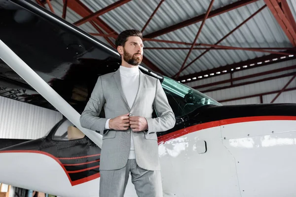 Vista basso angolo di uomo d'affari barbuto regolazione giacca e in piedi vicino elicottero moderno — Foto stock