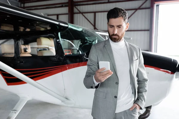 Hombre barbudo en ropa formal con teléfono inteligente y de pie con la mano en el bolsillo cerca de helicóptero moderno - foto de stock