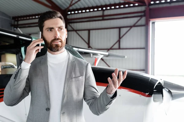 Бородатый мужчина в формальной одежде разговаривает на смартфоне и жестикулирует рядом с современным вертолетом — стоковое фото