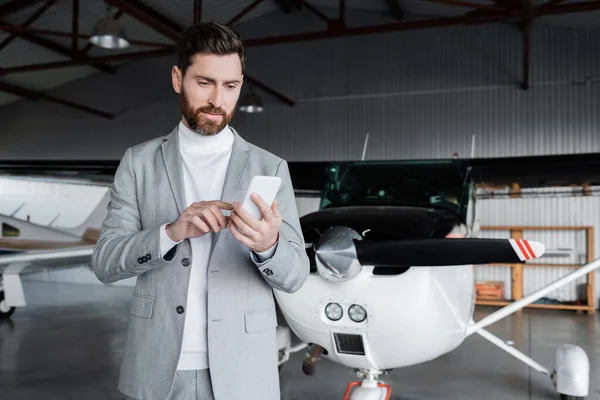 Hombre barbudo en traje usando teléfono inteligente cerca de helicóptero moderno - foto de stock