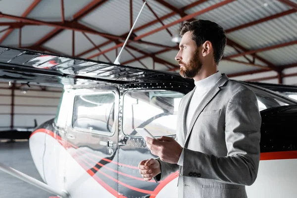 Hombre barbudo distraído en desgaste formal sosteniendo teléfono inteligente cerca de helicóptero moderno - foto de stock