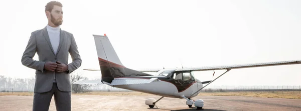 Homem de negócios barbudo ajustando blazer e de pé perto de helicóptero ao ar livre, bandeira — Fotografia de Stock