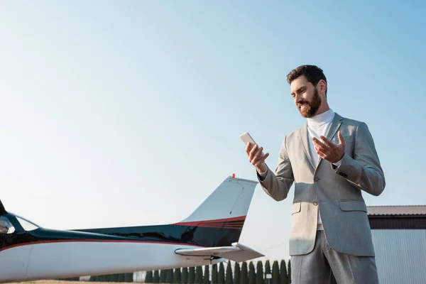 Bearded businessman smiling while looking at smartphone near helicopter outdoors — Stock Photo