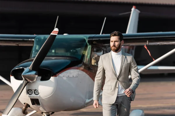 Homem de negócios barbudo em terno andando perto de helicóptero moderno e embaçado ao ar livre — Fotografia de Stock