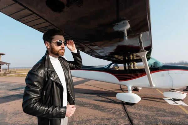 Bearded pilot in leather jacket adjusting sunglasses under aircraft wing — Stock Photo