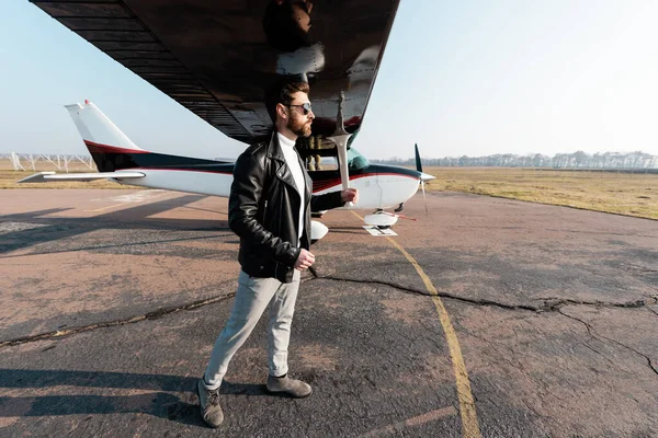 Comprimento total do piloto barbudo em óculos de sol e jaqueta de couro em pé sob asa da aeronave — Fotografia de Stock
