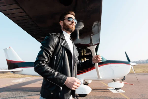 Vista a basso angolo del pilota barbuto in occhiali da sole e giacca di pelle in piedi sotto l'ala dell'aeromobile — Foto stock