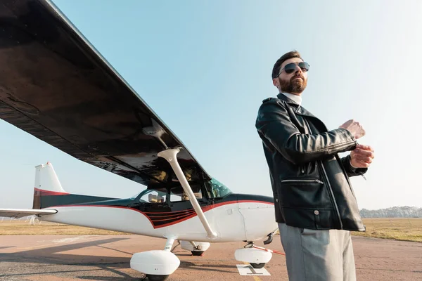 Vista de bajo ángulo del piloto en gafas de sol y chaqueta de cuero de pie cerca de la aeronave - foto de stock