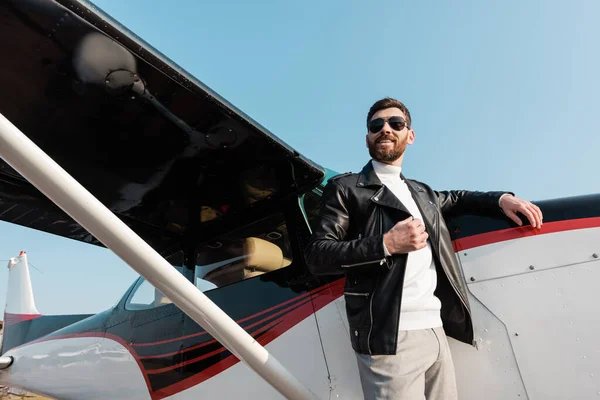 Vista de ángulo bajo de piloto feliz en chaqueta de cuero y gafas de sol de pie cerca de los aviones - foto de stock