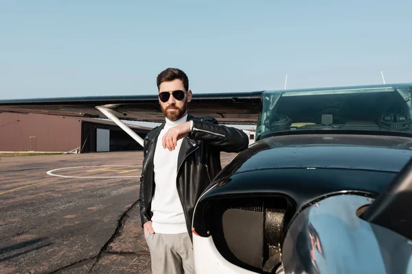 Bearded pilot in leather jacket and sunglasses standing with hand in pocket near aircraft — Stock Photo