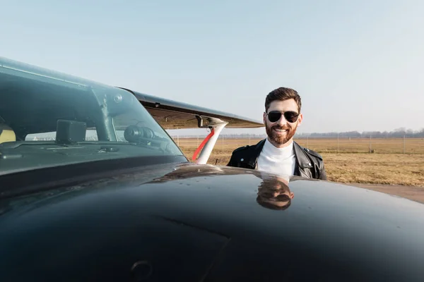 Smiling pilot in leather jacket and sunglasses near aircraft — Stock Photo