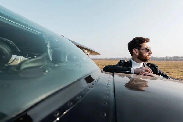 Piloto barbudo en chaqueta de cuero y gafas de sol mirando lejos cerca de los aviones - foto de stock