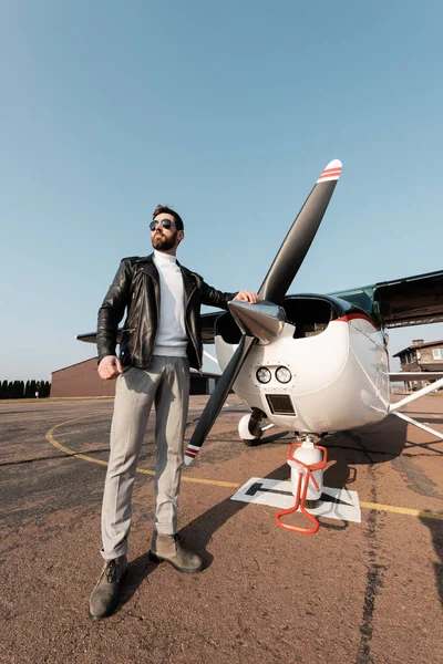 Longitud completa del piloto barbudo en chaqueta de cuero y gafas de sol de pie cerca de la aeronave - foto de stock